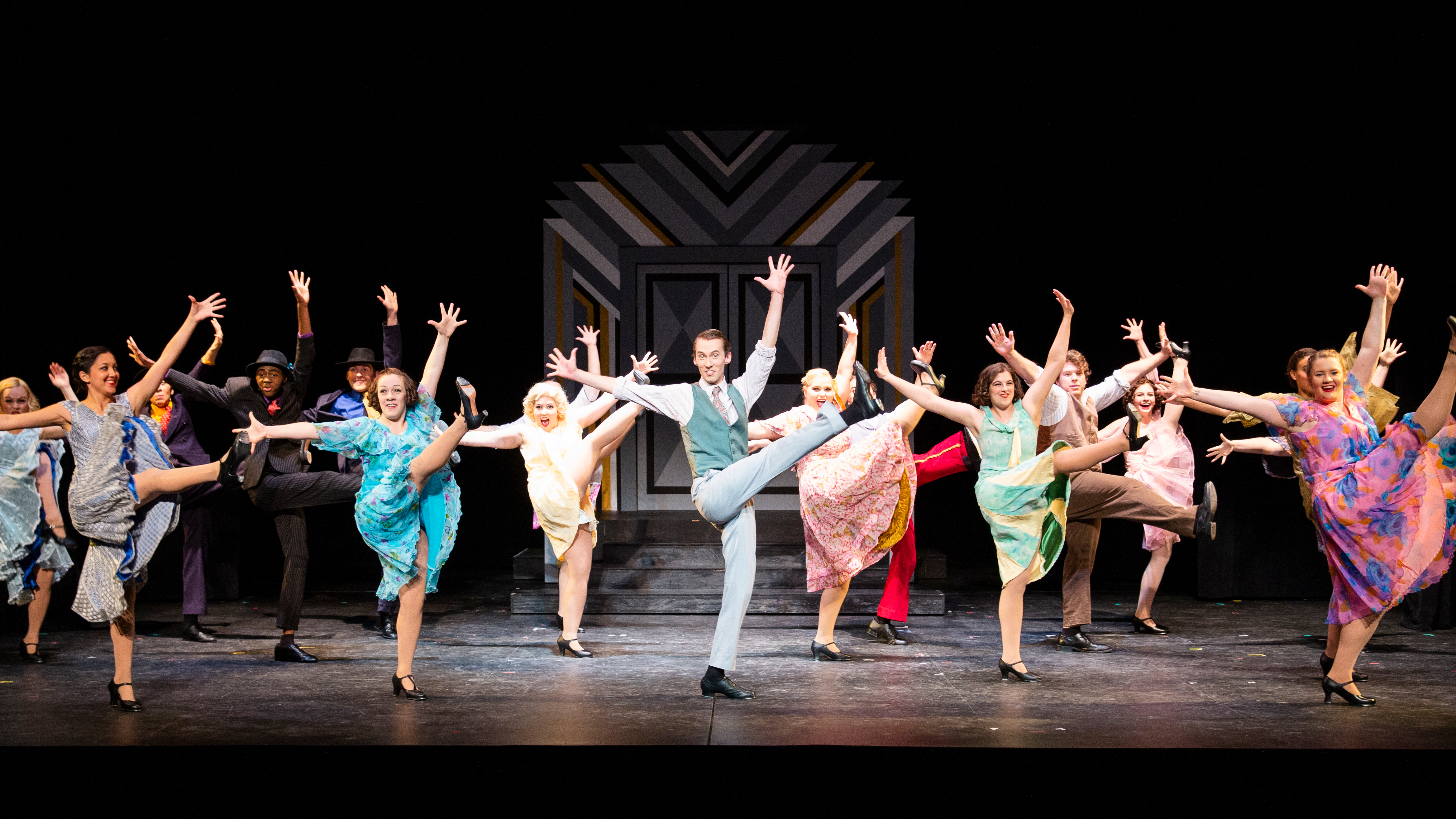 A chorus line of brightly dressed Drake Musical Theatre students raise their arms and legs during a production number