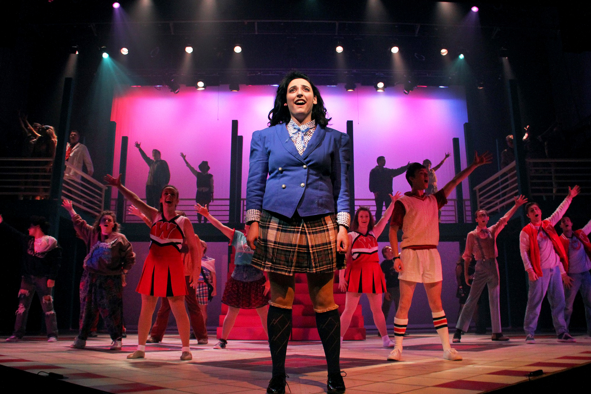 A Drake Musical Theatre student in a plaid skirt and double-breasted blazer sings a solo in front of a chorus for a local school production