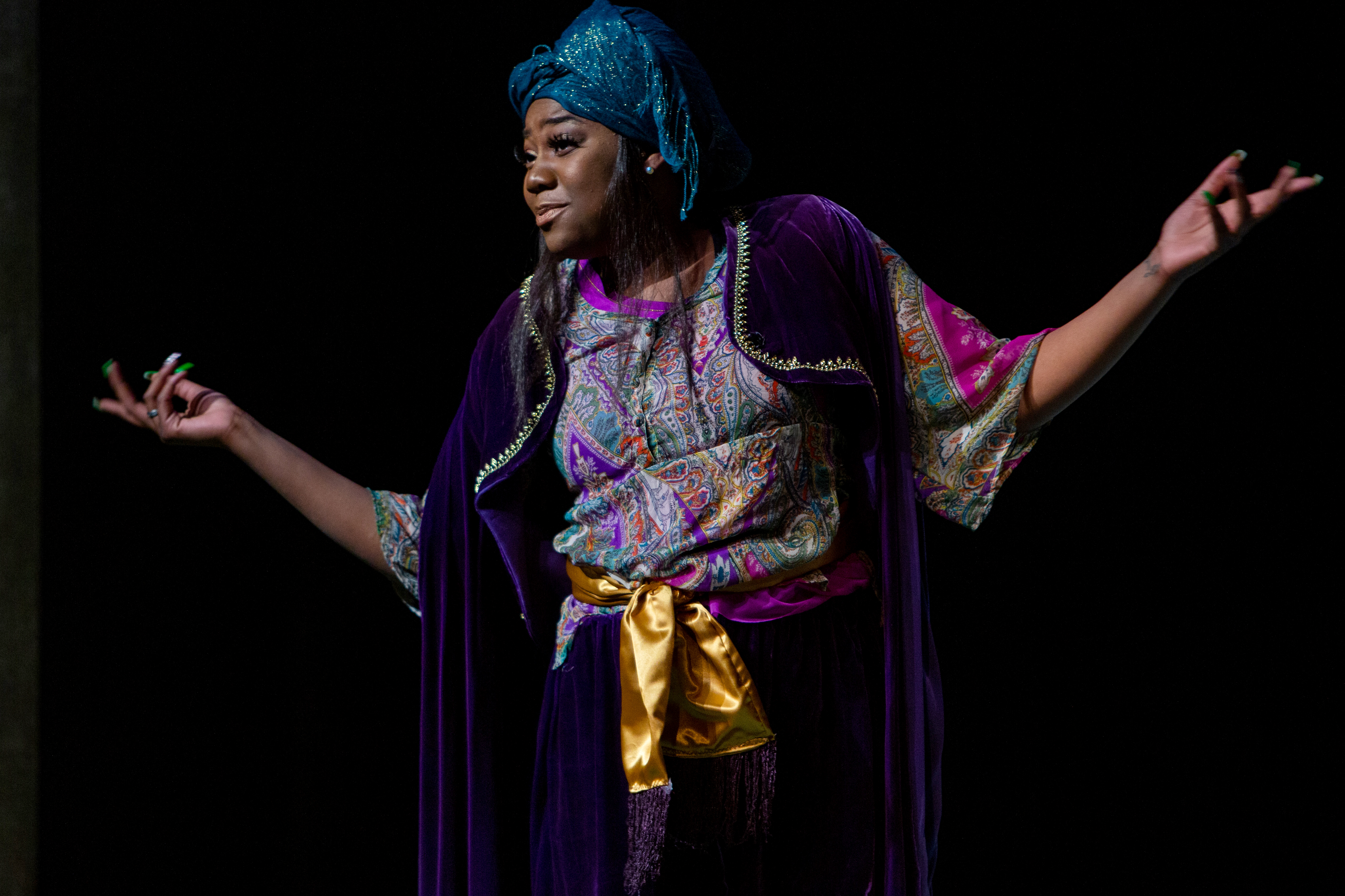 A Drake University Musical Theatre student wearing a silvery blue headwrap and purple velvet cape shrugs their shoulders and arms during a stage production