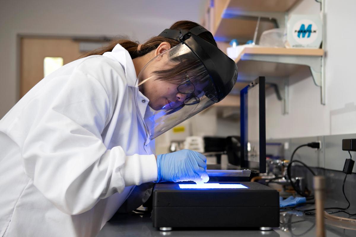 A Drake Neuroscience student, wearing a face mask, a lab coat, and gloves, examines a sample through a light box