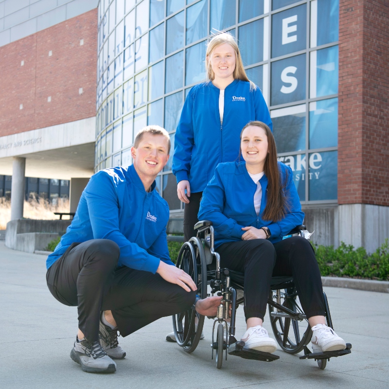 In front of a brick classroom building with curved windows, two Drake OTD students pose next to a classmate sitting in a wheelchair