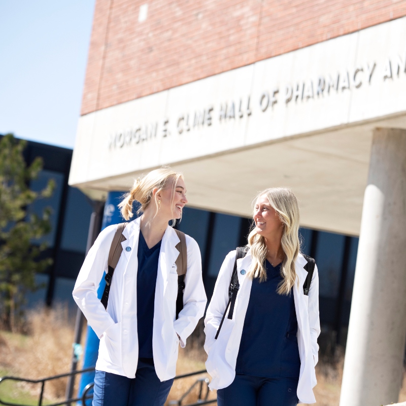 Two Drake PharmD students in lab coats and backpacks smile as they walk through campus on a sunny day
