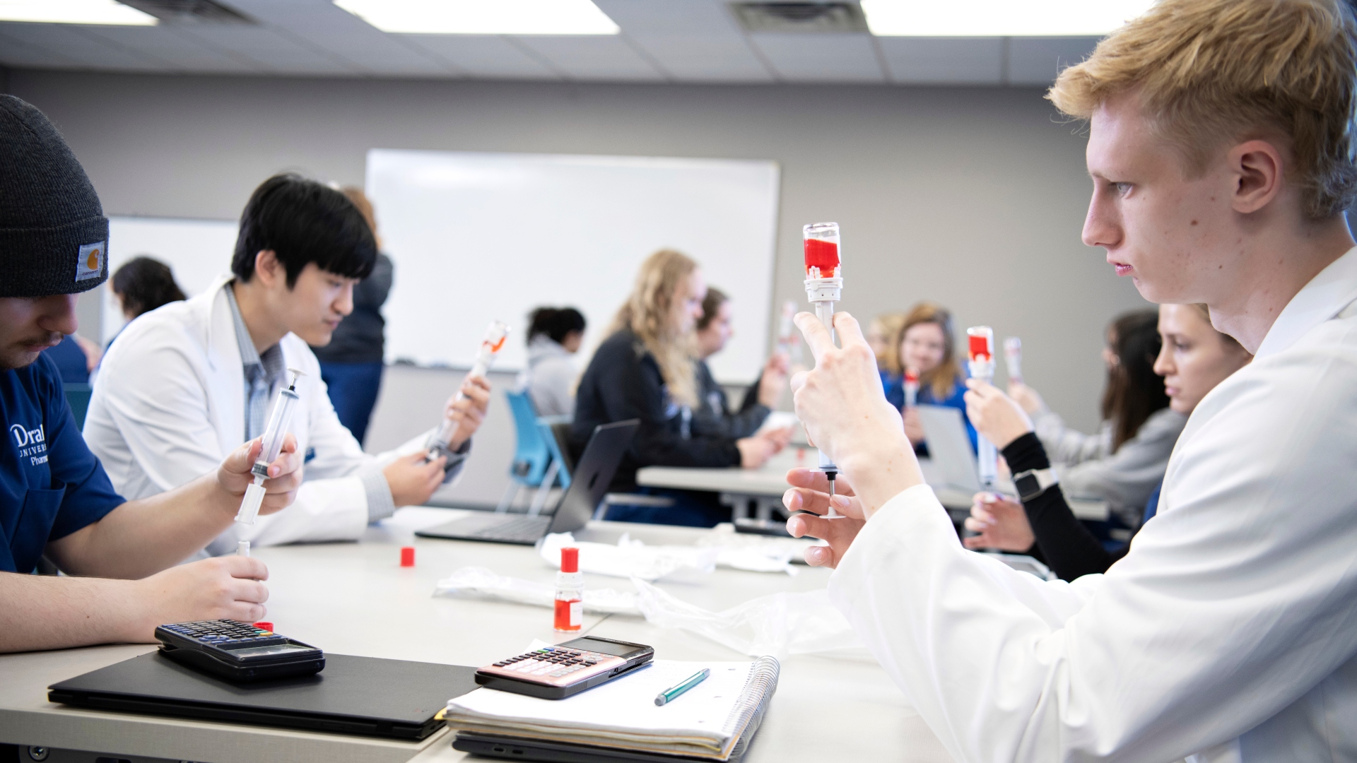 Drake PharmD students, sitting by desks with notebooks, laptops, and calculators, practice drawing medication from a vial