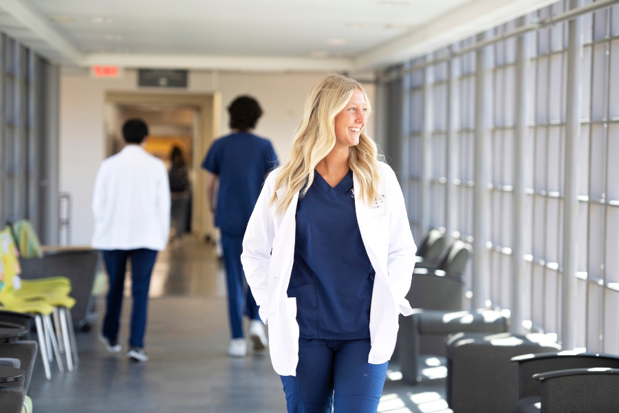 A Drake PharmD student, in blue scrubs and a white lab coat, walks through a hospital hallway