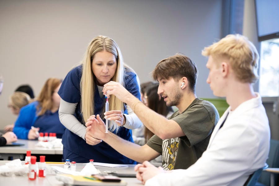 A Drake faculty member demonstrates how to draw medication with a syringe to two PharmD students sitting behind a desk