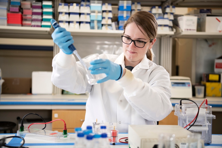 A Drake PharmD student in a lab coat, gloves, and safety glasses uses a syringe to draw liquid from a beaker