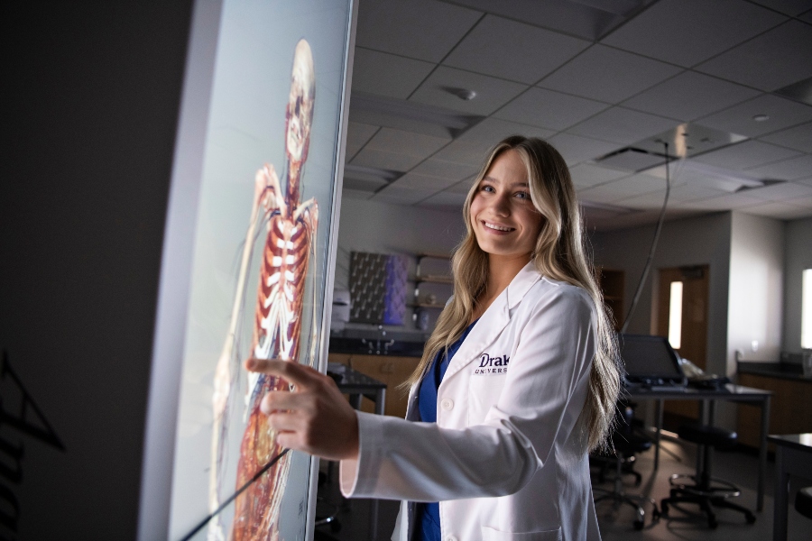 A Drake student in a white lab coat points to a lit anatomage table in a dark room