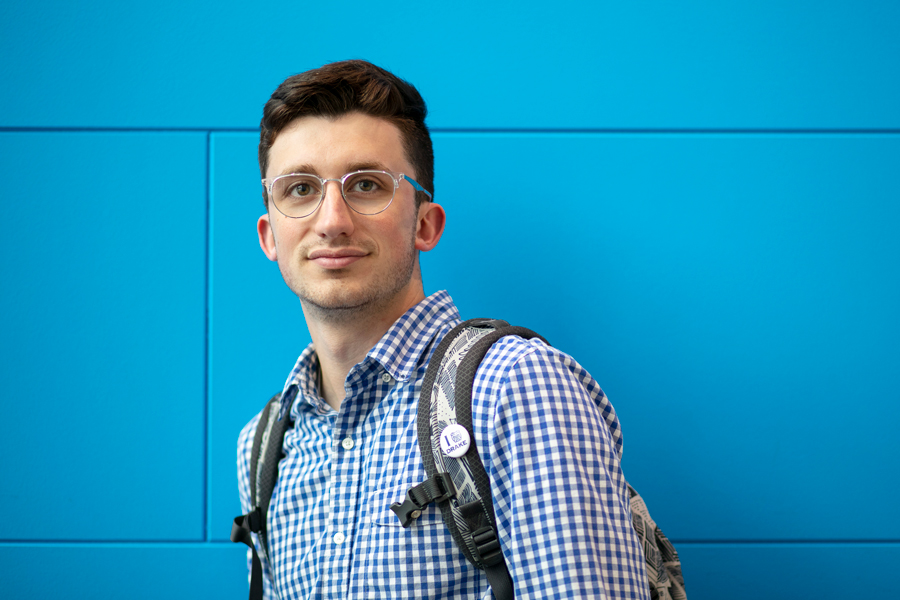 A Drake Physics students in glasses poses with a backpack in front of a blue wall