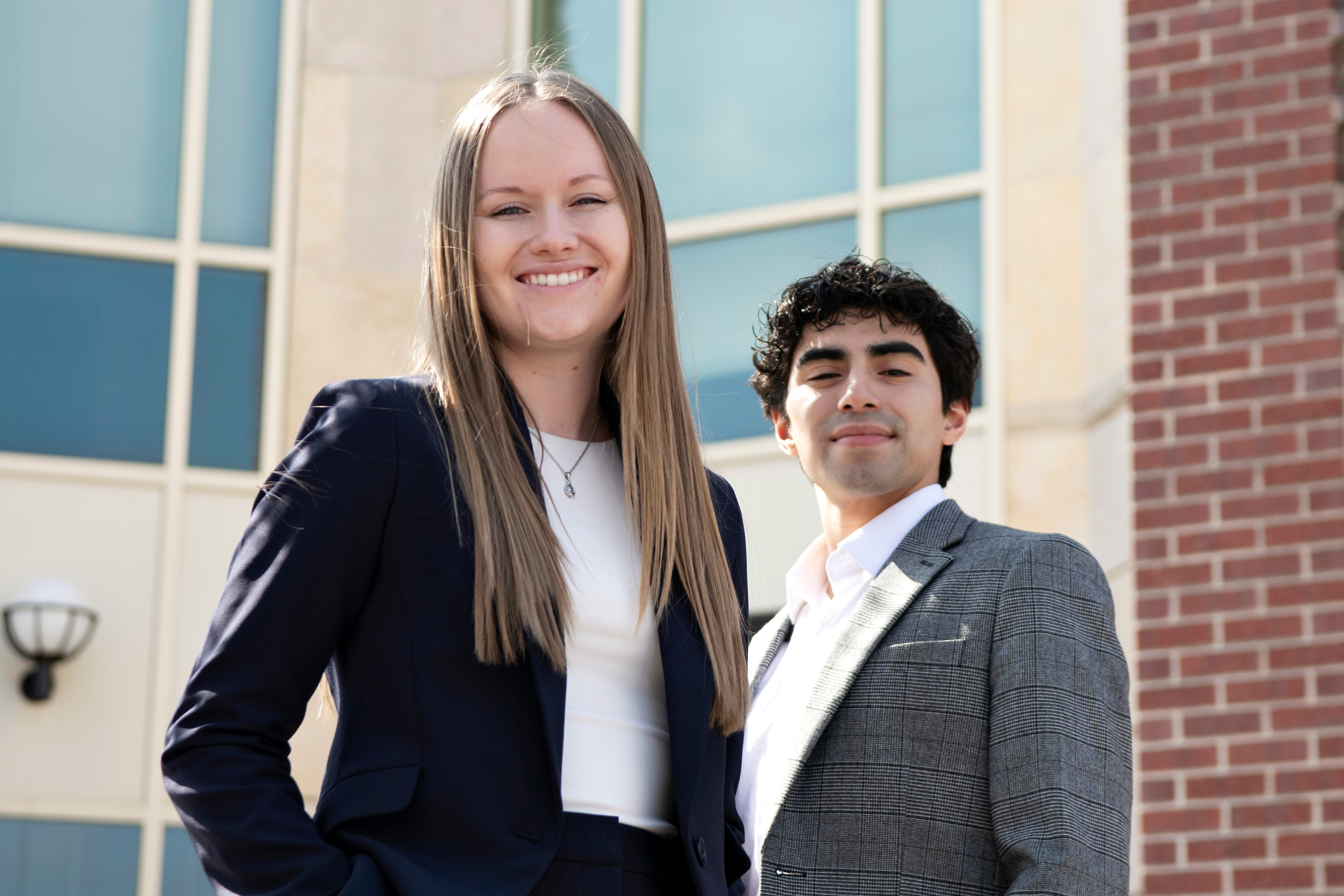 Two Drake Pre-Law students in suits stand in front of a red brick classroom building, one with their hand on their hip