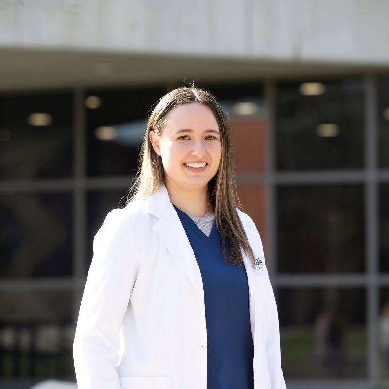 A Drake Pre-Medical student wearing a white lab coat and blue scrubs stands outside a campus building