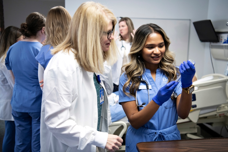 A faculty member instructs a Drake Pre-Medicine student, wearing scrubs and a stethoscope, in the correct usage of medical equipment