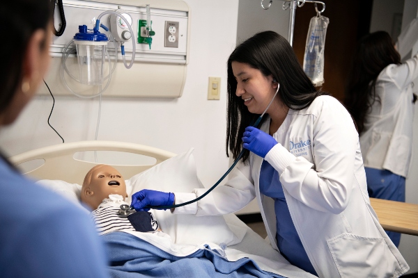 A Drake Pre-Nursing student in a white lab coat and gloves uses a stethoscope on a manikin