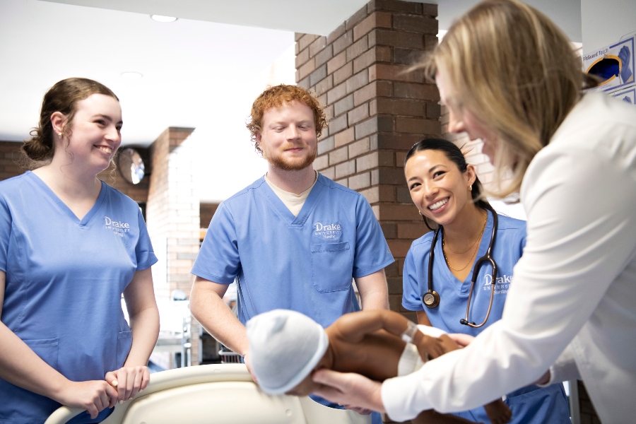Three Drake Pre-Nursing students in scrubs watch a faculty member's instruction involving an infant manikin