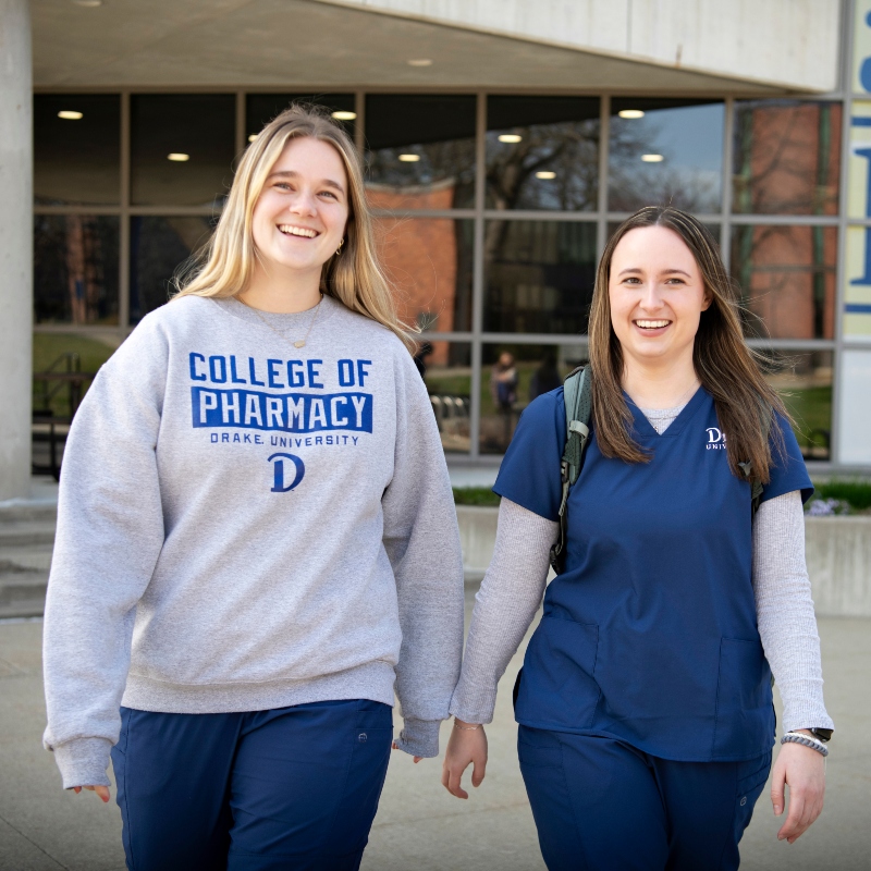 Two Drake Pre-Pharmacy students, one in a sweatshirt and another in scrubs, go for a walk outside the CPHS building
