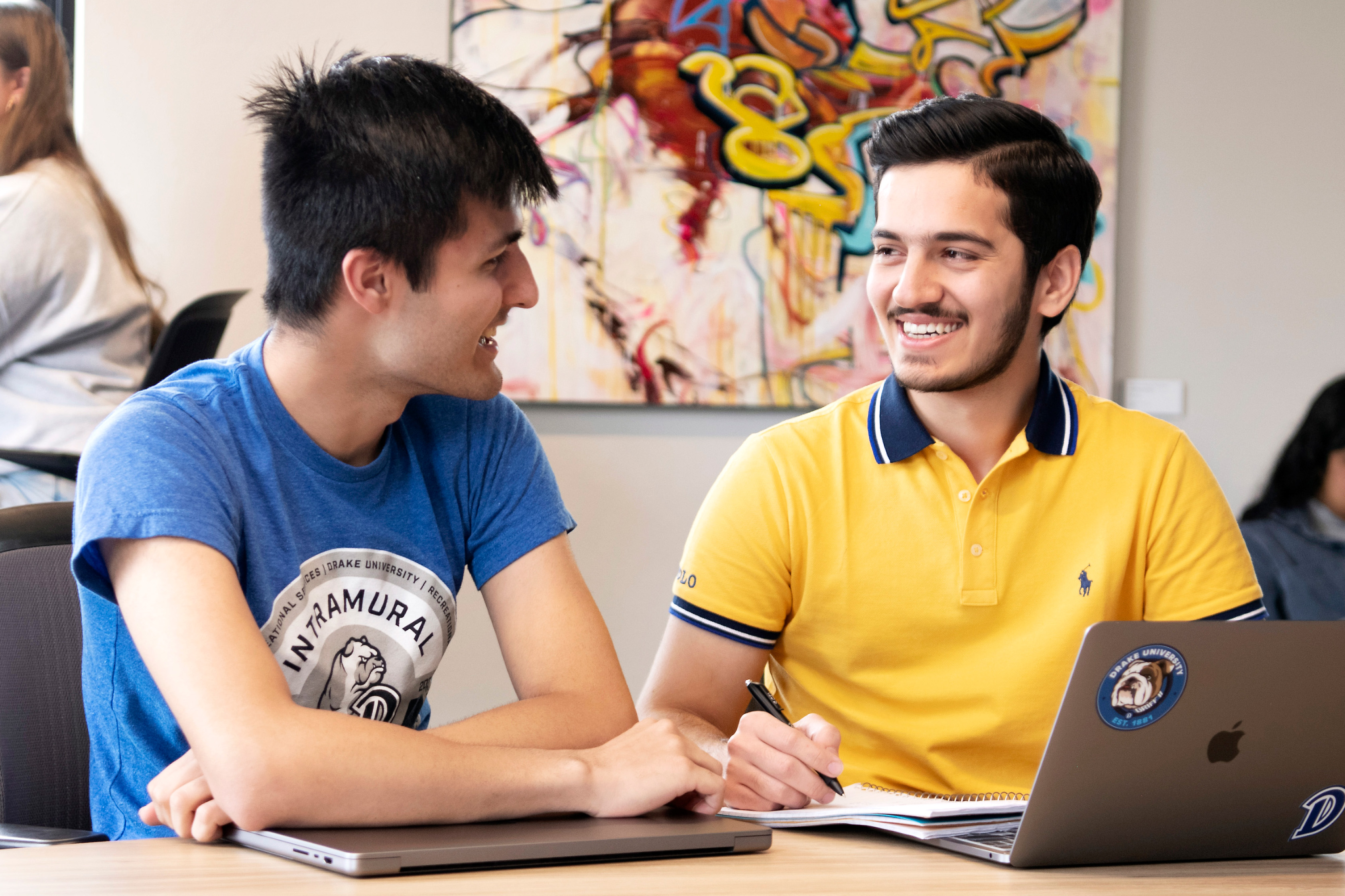 Two Drake Religious and Interfaith students converse behind a classroom desk with their laptops