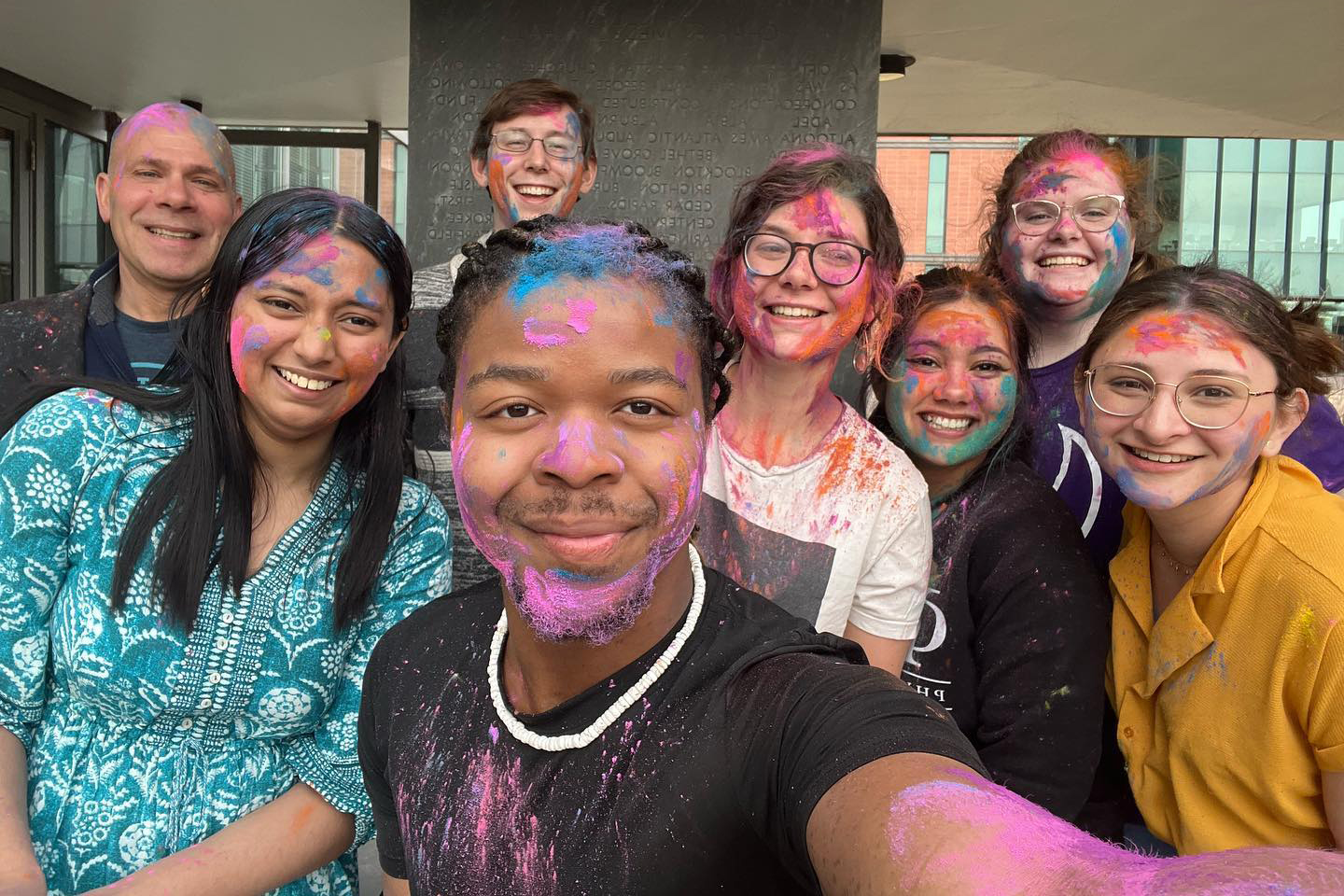 A group of Drake Religious and Interfaith Studies students, covered in brightly colored paint, pose in front of a building during a campus Holi celebration