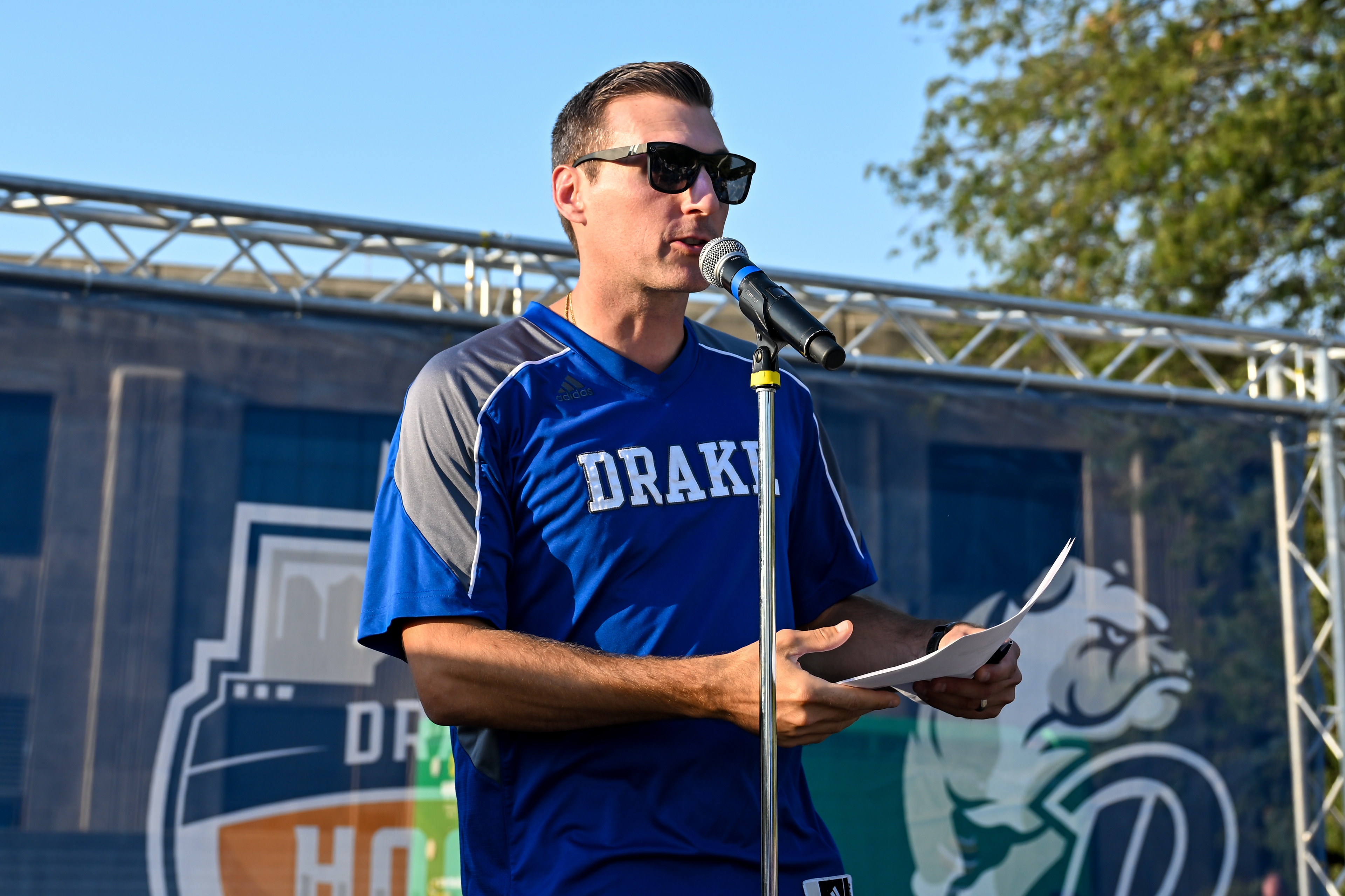 An alum in a Drake jersey stands on stage to present to a crowd of students outdoors