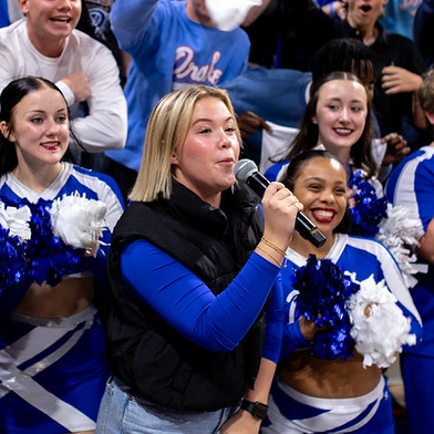 A student reporter from Drake's Sports Media and Communication program speaks into a microphone, with cheerleaders smiling and holding pom-poms in the background