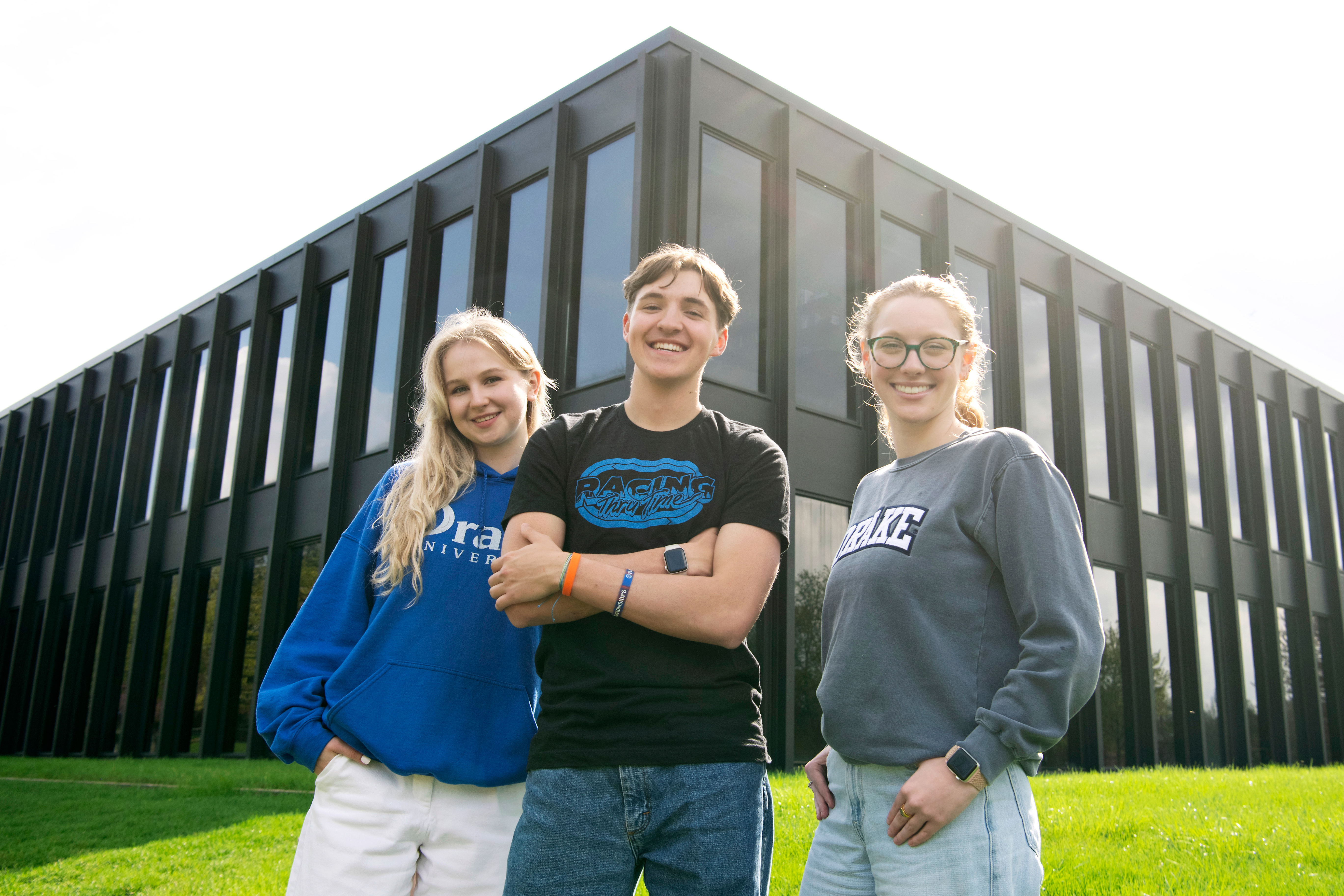 Three Drake students smile as they stand outdoors in front of a corner for the School of Journalism and Mass Communication building