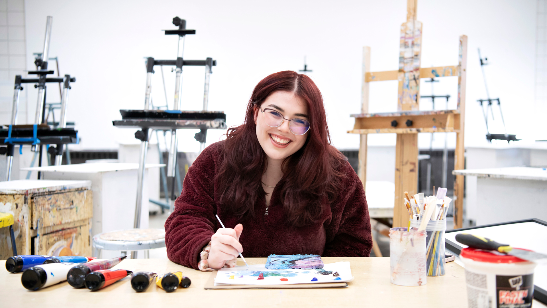 A student in glasses smiles while painting in one of Drake's art studios