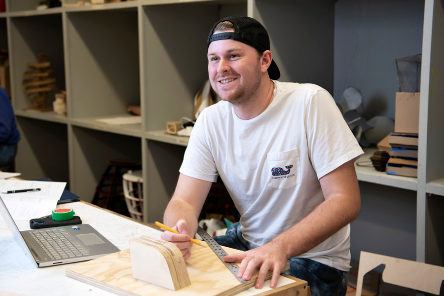 Sitting in front of a laptop, a Drake Studio Art student measures a piece of wood for a project
