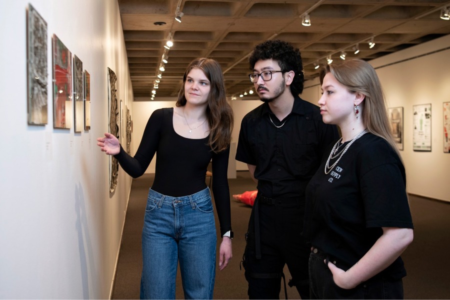 Three Drake Studio Art students discuss a triptych on the wall of a campus gallery