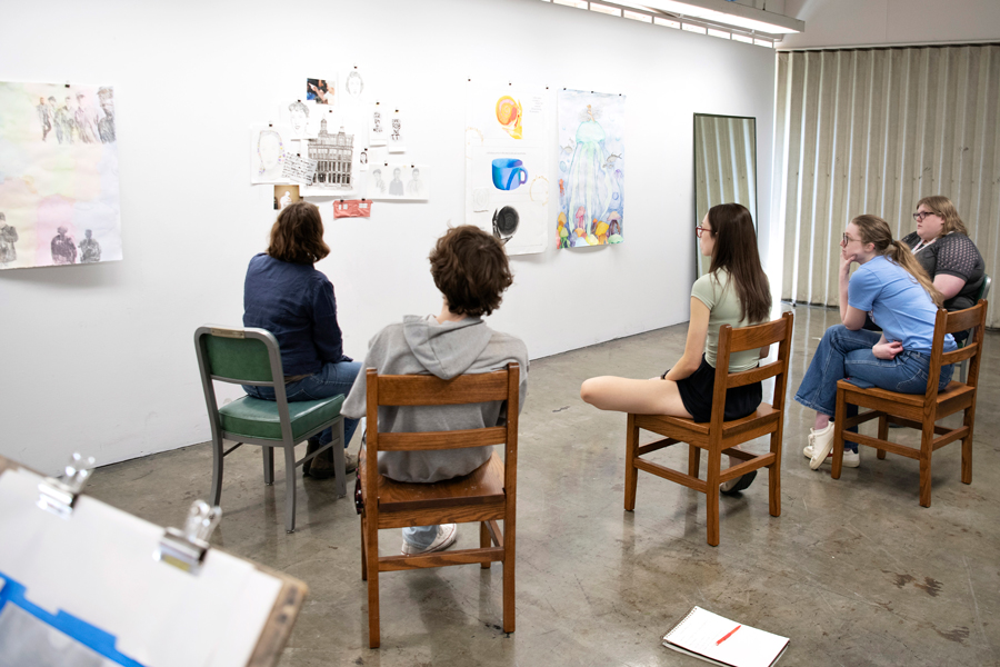 In a campus studio space, Studio Art students and a faculty member sitting in a half-circle discuss works posted onto a wall