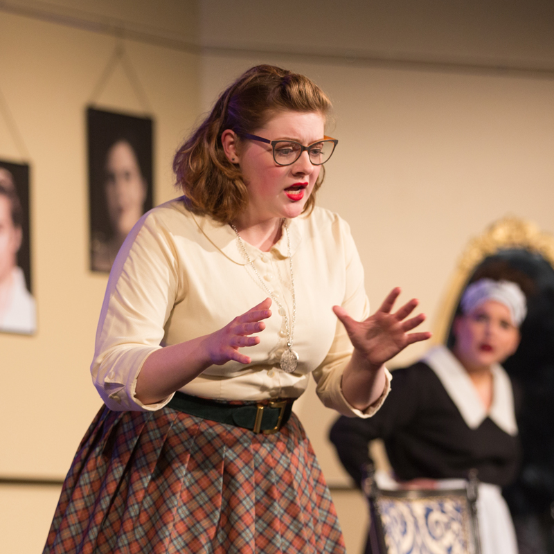 A Drake Theatre-Acting major gestures with their hands on a set with portraits in the background