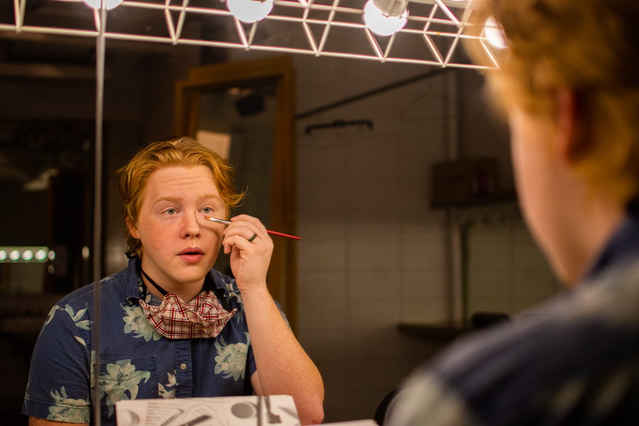 Looking into a mirror in a backstage dressing room, a Drake Theatre major applies makeup with a brush