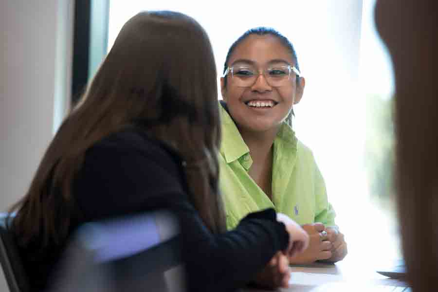 A student smiles at another student while sitting at a table