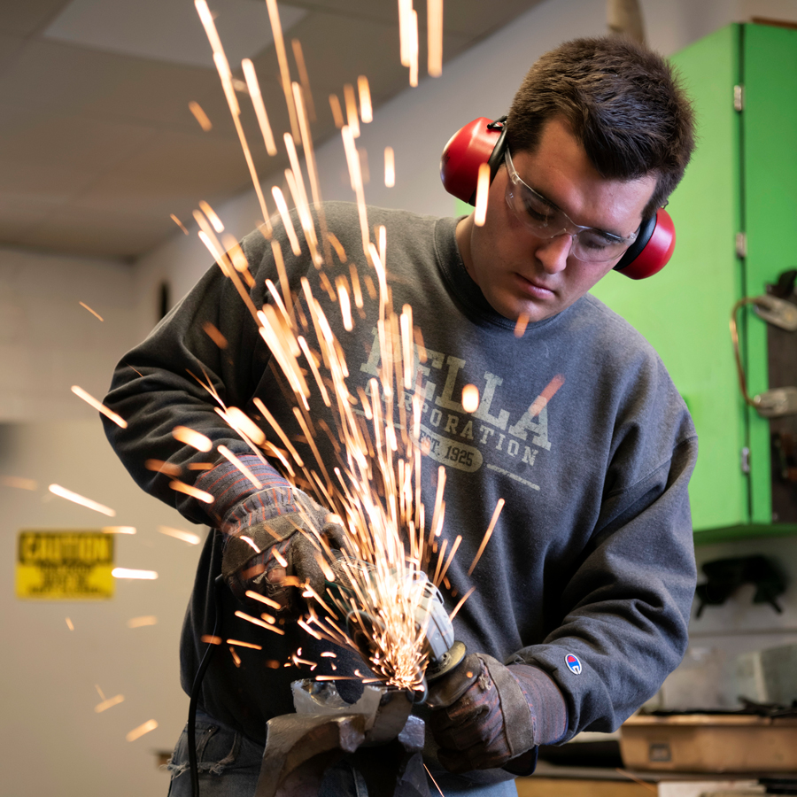 A student wearing protective earwear uses a metal grinder with sparks flying