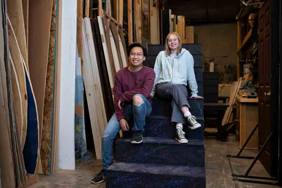 Two students smile while sitting on a staircase set piece backstage