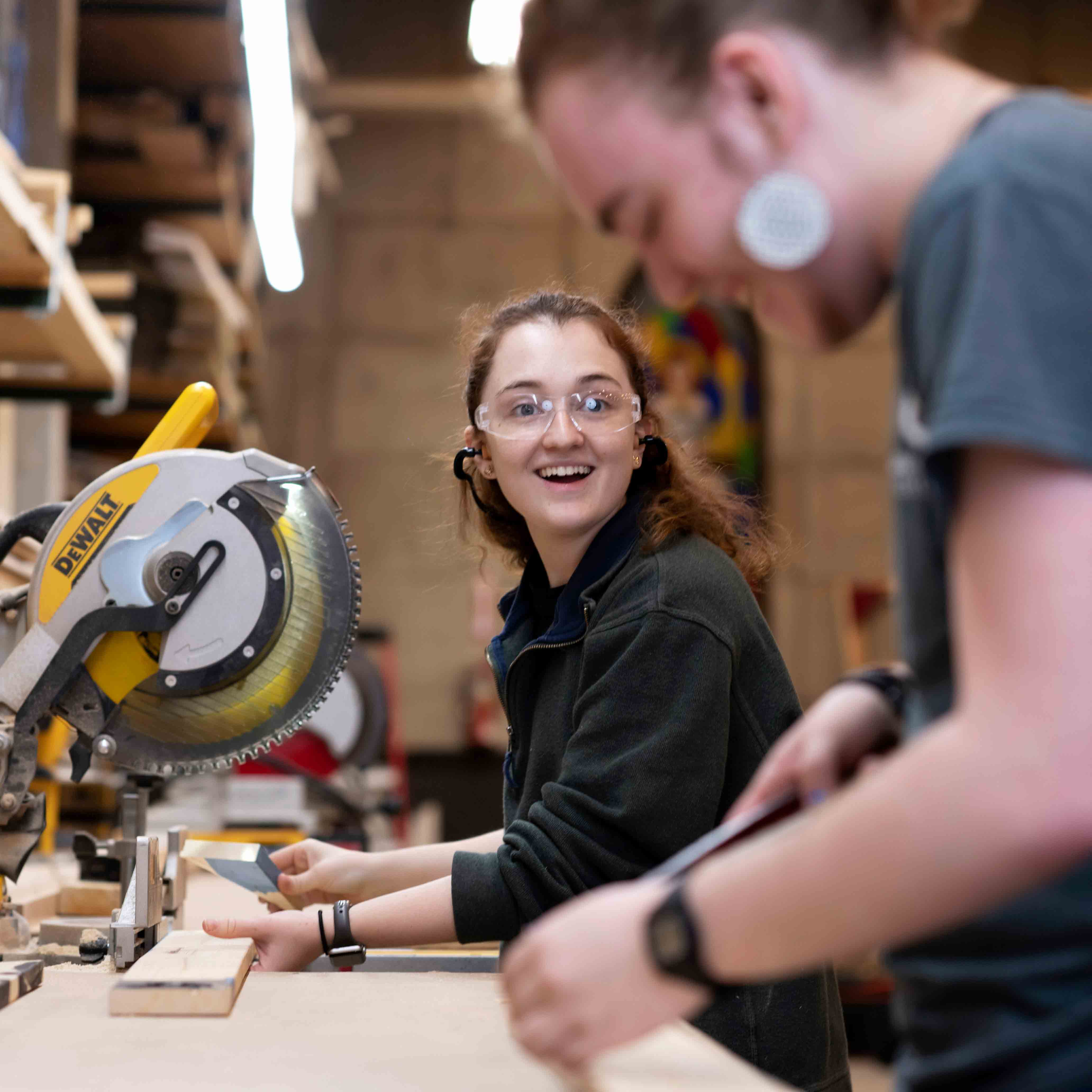 A student wearing safety goggles and operating a table saw smiles at another student