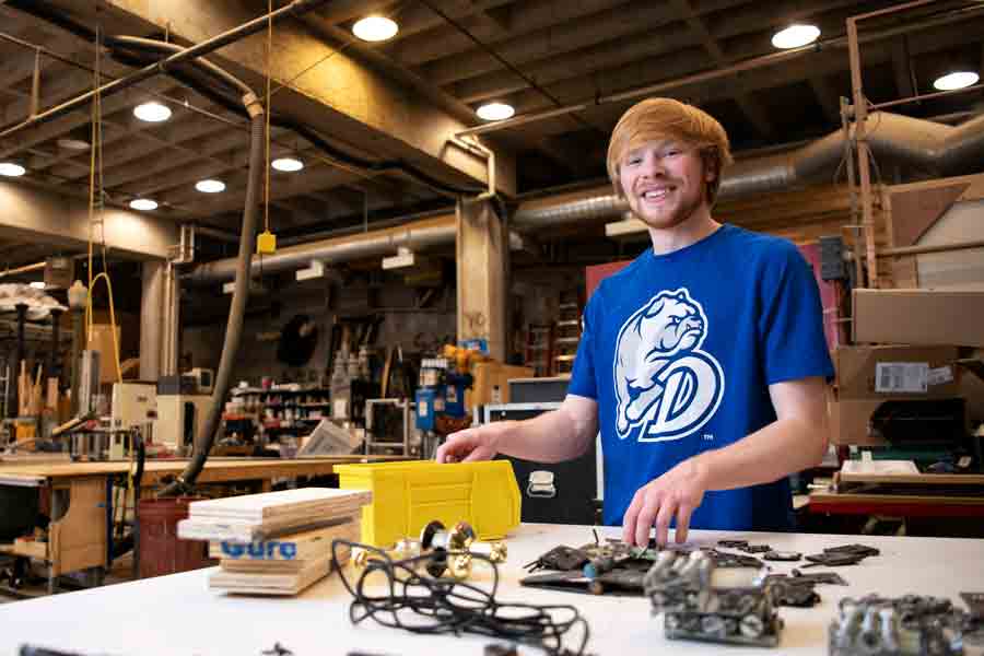 A student smiles at the camera standing in front of a table of design pieces