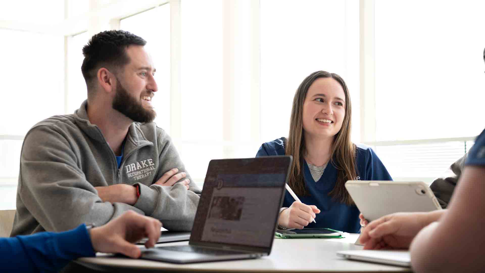 Two students sit at a table smiling and looking at other students off-camera