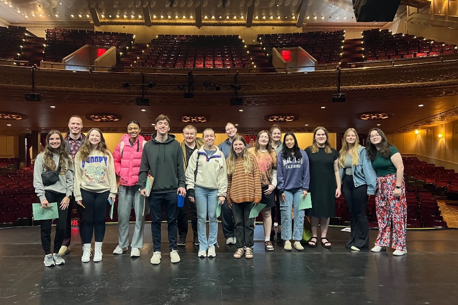 A class of students visiting a highschool posing on a stage and smiling