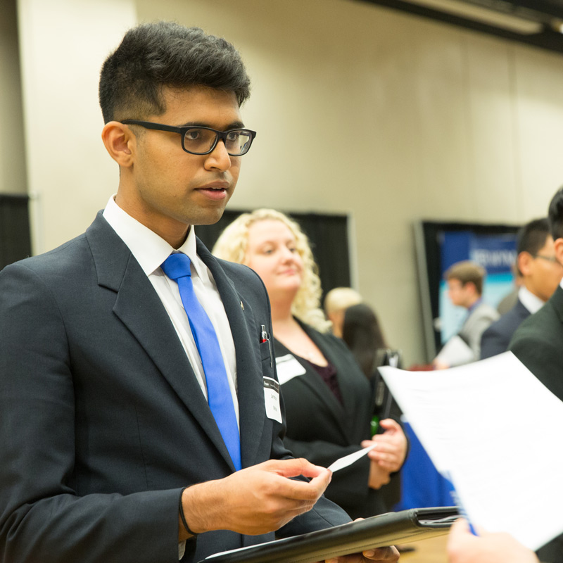 A student wearing glasses and a suit holding a padfolio speaking with a recruiter at a Drake University career fair