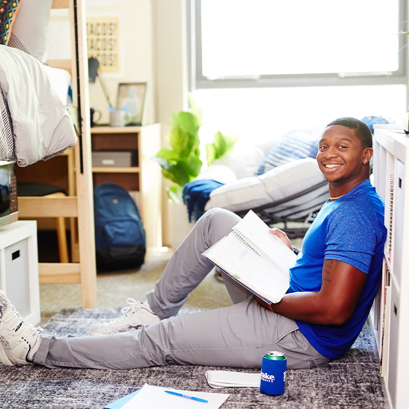 A Drake University student sitting on the floor of a dorm room holding a notebook and smiling