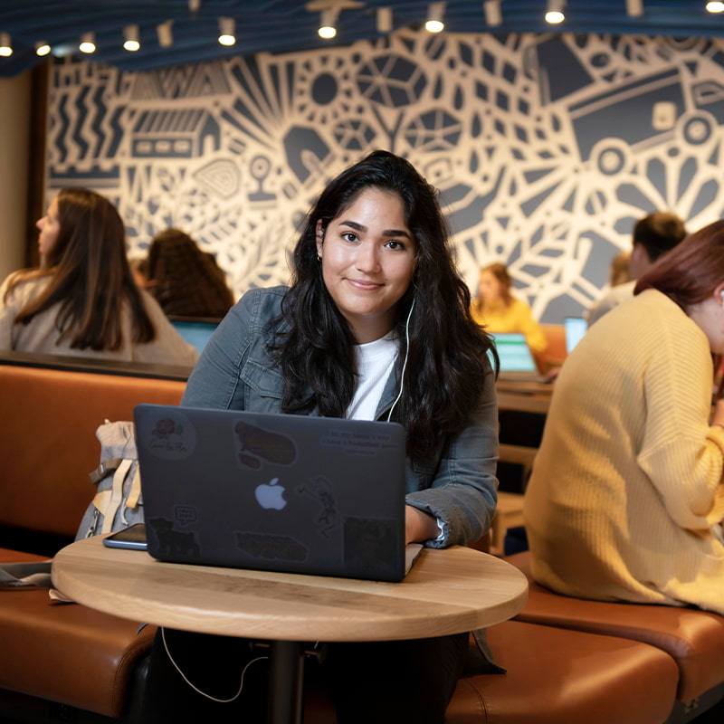 A Drake University student smiling while working on a laptop sitting at a single table in a dining location on campus