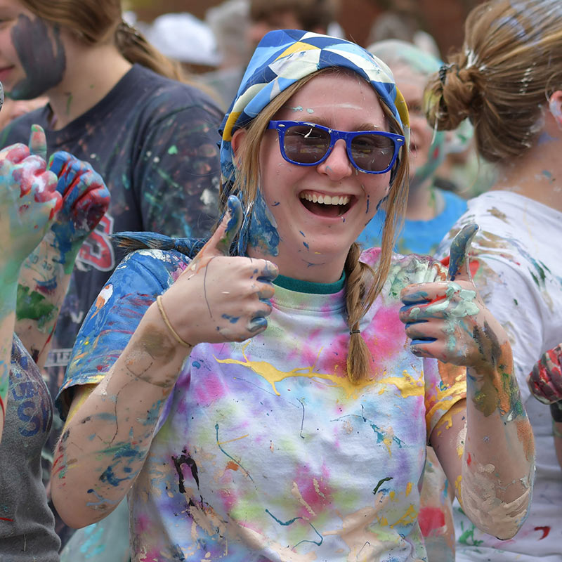 A student wearing sunglasses and a t-shirt covered in paint during the Drake Relays street painting event