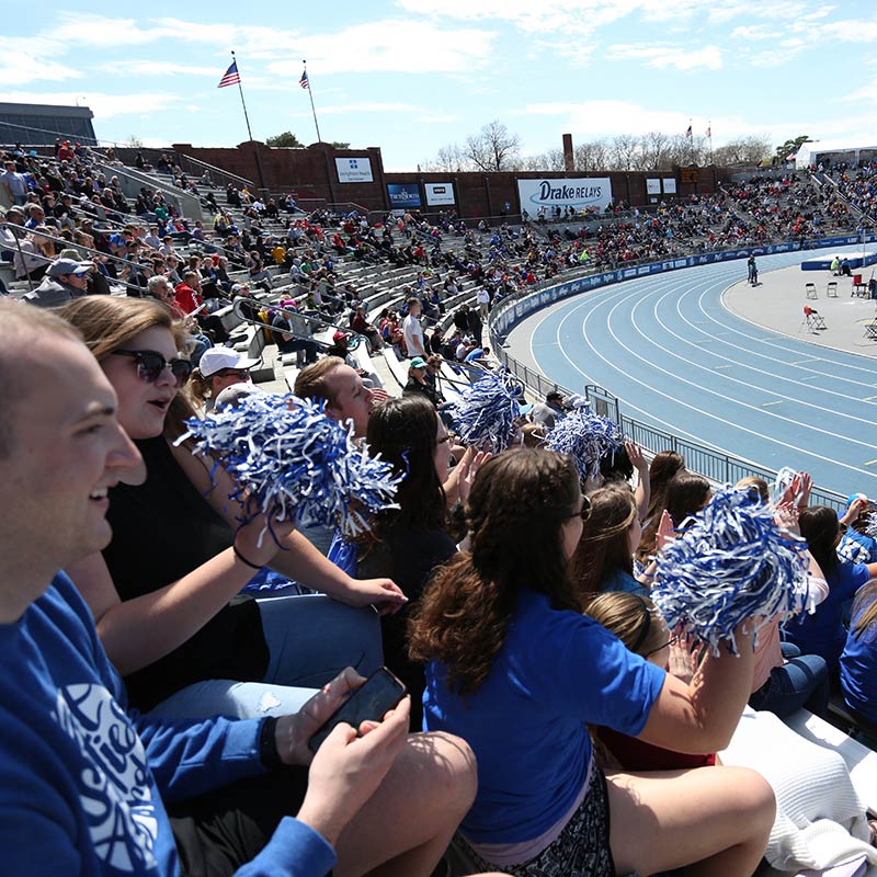 A crowd sitting in the bleachers of the blue oval track cheering during a Drake Relays event