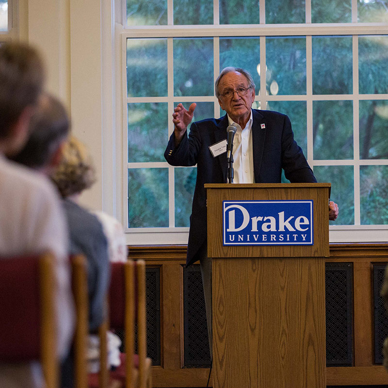 A guest speaker standing at a podium presenting to a group of Drake University students on campus