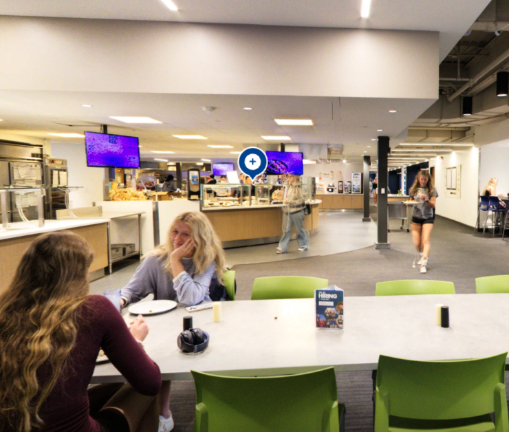 Students sit and chat at a table in a modern cafeteria with green chairs, while others walk near food counters and digital menu screens in the background.