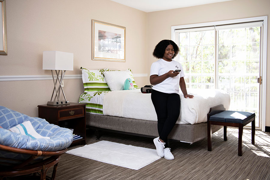 Person sitting on the edge of a neatly made bed, holding a phone in a bright bedroom with a large sliding glass door.