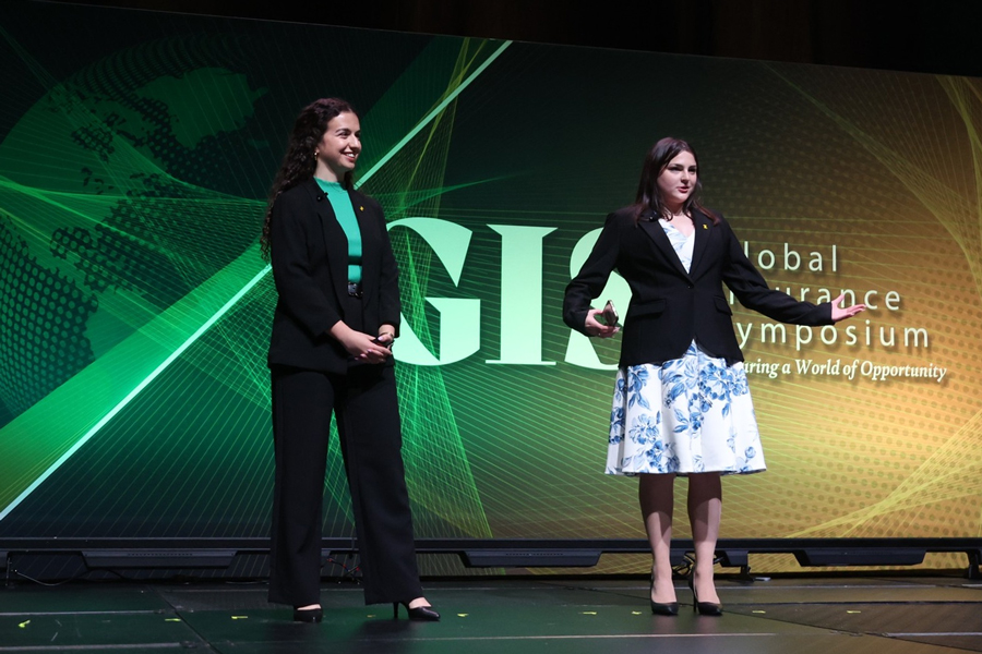 Two woman in business attire stand on stage with a presentation for the Global Insurance Symposium in background